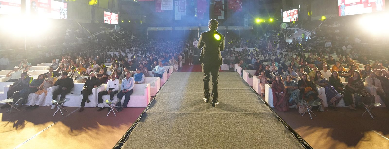 A facilitator addressing a large corporate audience on a runway stage